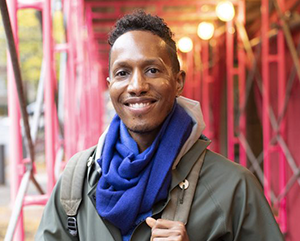 Portrait of Uri McMillan wearing a green jacket and blue scarf, standing outside beneath a red protective scaffolding.
