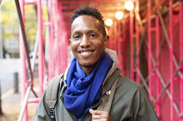 Portrait of Uri McMillan wearing a green jacket and blue scarf, standing outside beneath a red protective scaffolding.