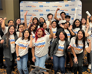 A group of students in UCLA College Corps shirts cheer in front of a step-and-repeat branded with UCLA College Corps logos.