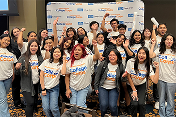 A group of students in UCLA College Corps shirts cheer in front of a step-and-repeat branded with UCLA College Corps logos.