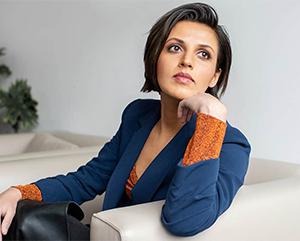 Prineha Narang wearing a blue coat over an orange sweater in a room with white walls and furniture.