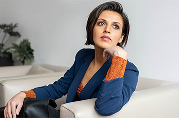Prineha Narang wearing a blue coat over an orange sweater in a room with white walls and furniture.