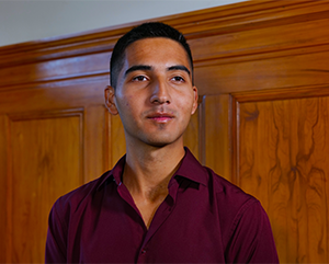 Truman Scholar_Diego Sarmiento_363-241 A portrait of Diego Sarmiento in a maroon dress shirt with a stained wood wall in the background.