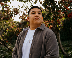 Nilton Serva wearing a light brown sweater and a white shirt, standing outside with trees, bushes and sunlight in the background.