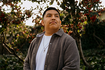 Nilton Serva wearing a light brown sweater and a white shirt, standing outside with trees, bushes and sunlight in the background.