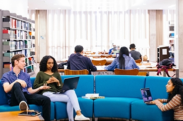 Student sitting inside a well lit room with a blue couch and communal desks in the background.