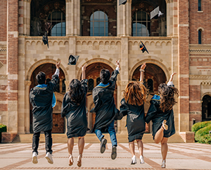2024-COMMENCEMENT-363-241 A group of jubilant graduates in traditional black caps and gowns are captured mid-jump against a backdrop of Royce Hall with it's famed arched entryways. The graduates, facing away from the camera, are seen tossing their caps high into the air, marking a celebratory moment under the clear, sunny skies that signify the bright future ahead.