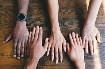 Five hands with different skin tones on a wooden table view from above.