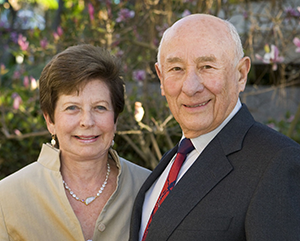 Luskins-FINAL-363-241 Renee (left) and Meyer Luskin standing outside on a sunny day with green leaves, branches and pink flowers in the background.