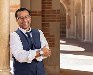 Paco+Retana-363-241 Paco Retana in a white dress shirt with a blue vest and tie standing in a stone corridor with silhouettes of columns in the background.
