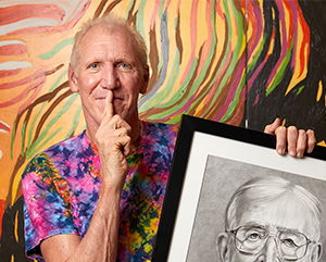 bill-walton_2-363-241 Bill Walton wearing a tie die shirt with his right index finger pressed to this lips while holding a black and white drawing of John Wooden against a painted wall with orange, yellow, red, green, brown and black streaks of paint.