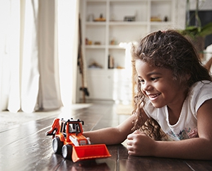Young Hispanic girl lying on the floor in the sitting room playing with a toy digger truck A young girl lying on floor playing with an orange bulldozer.