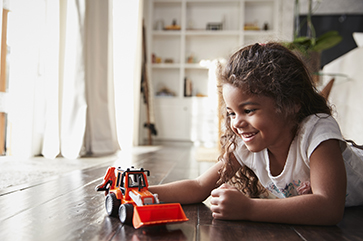 A young girl lying on floor playing with an orange bulldozer.