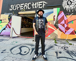 Erick+Superchief-363-241 (1) Erick Zerecero, dressed in black, standing in front of Superchief sign