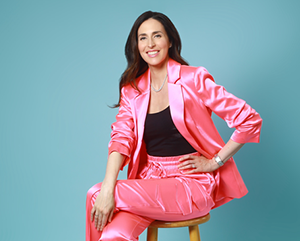 Hannah-Gordon-in-pink-343-241 Hannah Gordon is a pink suit with a black top, sitting on a wooden barstool in front of a light blue photo studio background.