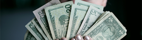 A close-up image of a blue-eyed blonde-haired woman holding a fan of U.S. dollars