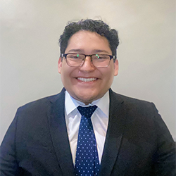 Gustavo Calva Hernandez in a blue suit and tie, posing against a white wall. 