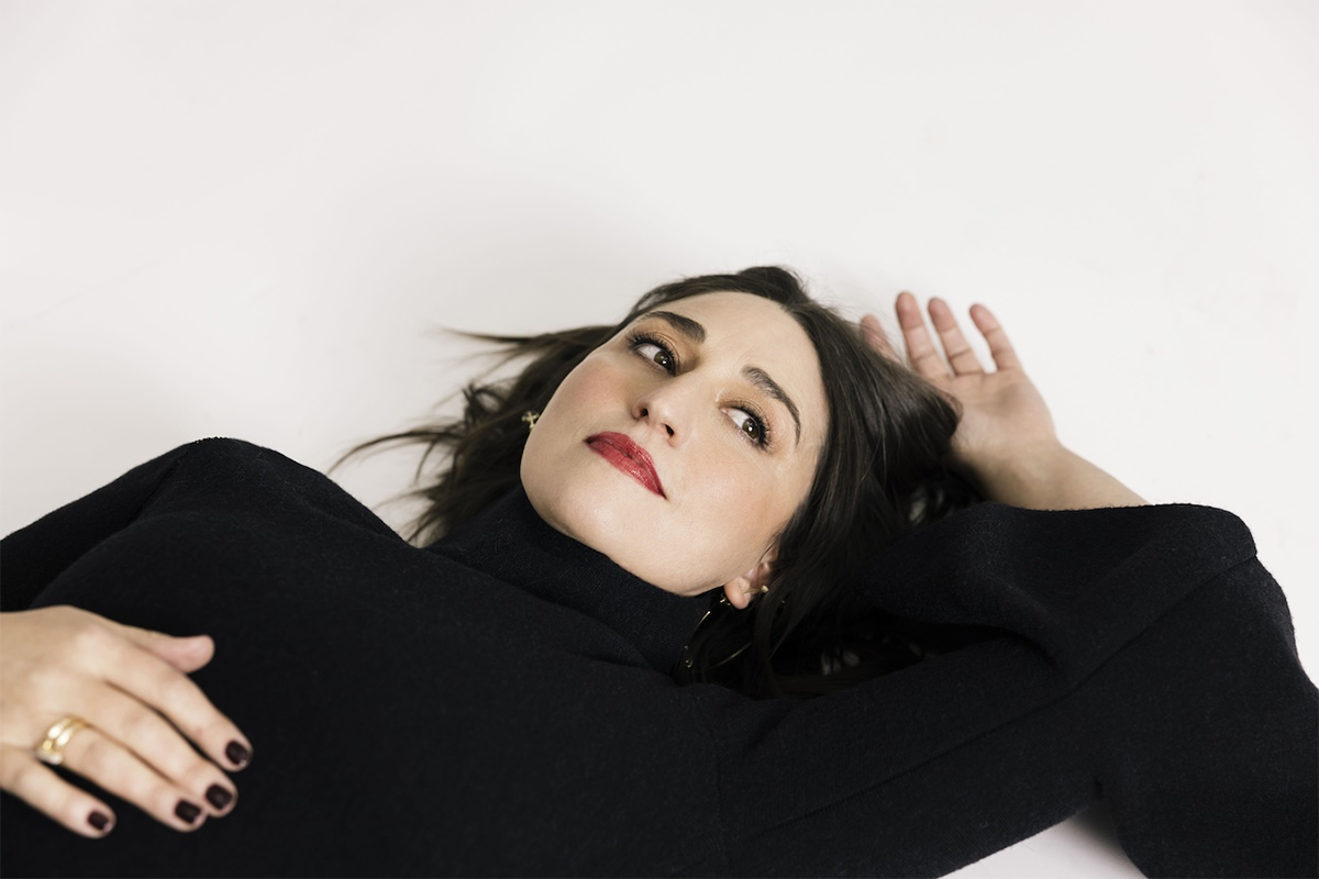 Sarah Bareilles in black lying on a white floor with left hand above her head