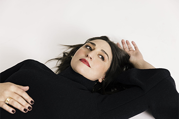 Sarah Bareilles in black lying on a white floor with left hand above her head