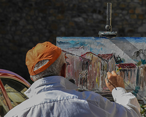Curiosity-363-241 Older man in red hat paints a picture of a building on a canvas sitting on an easel