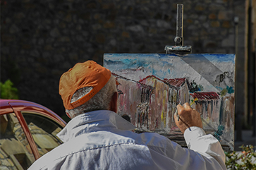 Older man in red hat paints a picture of a building on a canvas sitting on an easel