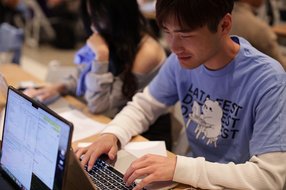 a student (wearinga DataFest tshirt) working on their laptop at a table with other students