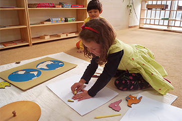 Preschool-aged girl in yellow dress plays on the floor with geography shape puzzle as young boy watches behind her