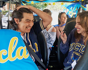 BGD+Gift+Cart-363-241 Four students smile as they sit in a golf cart with UCLA script logo covering the front seat