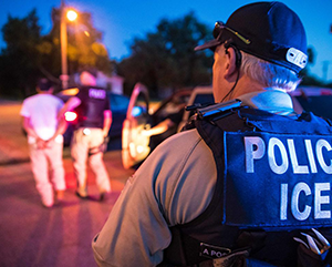 ICE-363-241 A man is seen from the back wearing a police ICE vest. On the left, two male figures can be seen in soft focus. One man has his hands behind his back, the other, who is wearing a police vest, is leading him