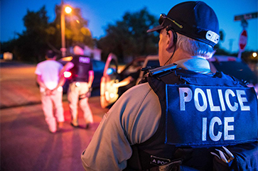A man is seen from the back wearing a police ICE vest. On the left, two male figures can be seen in soft focus. One man has his hands behind his back, the other, who is wearing a police vest, is leading him