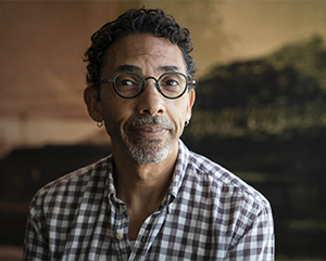 Robin-Kelley-headshot-2019-363-241 Robin D.G. Kelley in a white and grey checkered shirt against a painting of a sunset