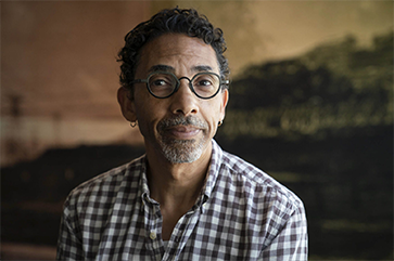 Robin D.G. Kelley in a white and grey checkered shirt against a painting of a sunset