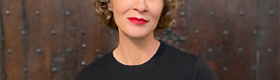 Tamar Christensen in a black sweater and striking red lipstick again a brown wood wall studded with carriage bolts.