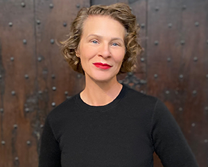 Tamar-Christensen-portrait-363-241 Tamar Christensen in a black sweater and striking red lipstick again a brown wood wall studded with carriage bolts.