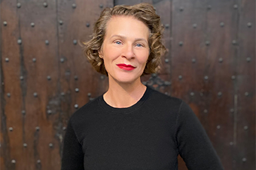 Tamar Christensen in a black sweater and striking red lipstick again a brown wood wall studded with carriage bolts.
