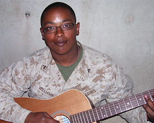 Jason-Williams-363-241 Jason Williams in military fatigues, leaning against a concrete wall whilst holding an acoustic guitar.