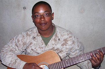 Jason Williams in military fatigues, leaning against a concrete wall whilst holding an acoustic guitar.