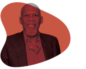 KAJ-363-241 A man with a trimmed beard and bald head, wearing a dark blazer over a red shirt, smiles against a solid red abstract background shape.