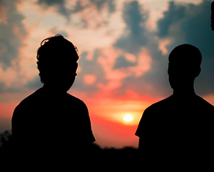 sunset-6269536_1280-363-241 Silhouettes of two people standing outdoors against a dramatic sunset sky, with vibrant hues of orange, red, and blue and scattered clouds.