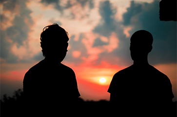 Silhouettes of two people standing outdoors against a dramatic sunset sky, with vibrant hues of orange, red, and blue and scattered clouds.
