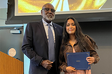 Charles Alexander in a dark suit and blue tie stands next to a student in brown jacket and peach pants in front of an overhead projector screen. 