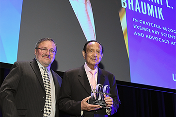 Miguel García-Garibay, left, in black jacket and diamond-patterned tie, and Mani Bhaumik, in black jacket and pink tie, holding a UCLA College 100 trophy