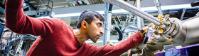 A grad student focuses intently as he slides a three-foot-long steel bar into a large apparatus made of tubes and wires, in a lab full of similar such apparatuses.