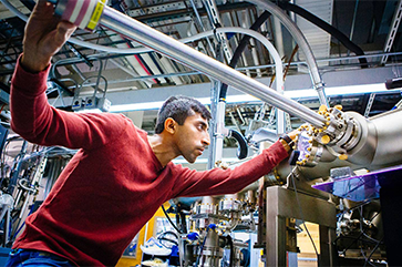A grad student focuses intently as he slides a three-foot-long steel bar into a large apparatus made of tubes and wires, in a lab full of similar such apparatuses.