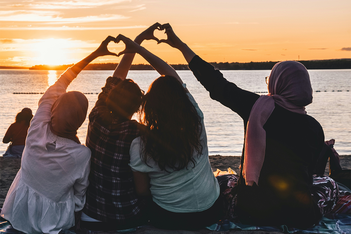 Four people sit closely together on a beach blanket, facing a body of water at sunset. Their arms are raised, forming heart shapes with their hands against the glowing orange sky.