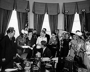 Black and white photo of a group of women and men around a desk