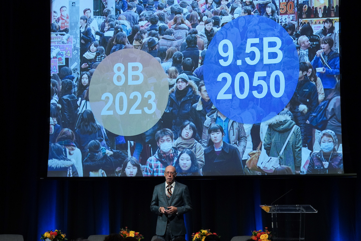 Jack Bobo delivering a keynote speech. Behind him is a large screen showing a densely crowded street filled with people. Two large circles are superimposed on the image: an orange circle on the left with the text "8B 2023" and a blue circle on the right with the text "9.5B 2050," indicating global population figures for those years.