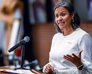 Amada Armenta, in white, gestures with her left hand as she speaks at a podium