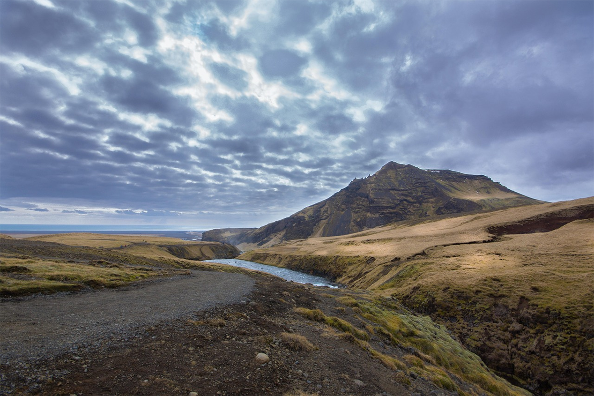 A dramatic mountain landscape under a moody sky. A sharp, jagged peak rises in the distance, partially shrouded by dense, textured clouds. In the foreground, a gravel path curves gently through a valley of rocky terrain and dry grass, with patches of moss adding muted green tones. A winding river cuts through the middle ground, reflecting the filtered light that breaks through the clouds.
