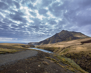 A dramatic mountain landscape under a moody sky. A sharp, jagged peak rises in the distance, partially shrouded by dense, textured clouds. In the foreground, a gravel path curves gently through a valley of rocky terrain and dry grass, with patches of moss adding muted green tones. A winding river cuts through the middle ground, reflecting the filtered light that breaks through the clouds.
